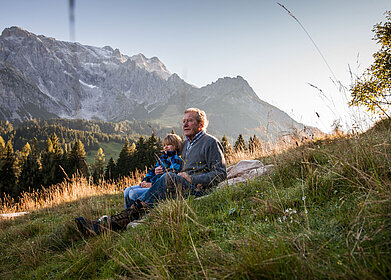 Großvater sitzt mit einem Kind auf einer Wiese und blickt in die Berglandschaft des Hochkönigs bei warmem Abendlicht.